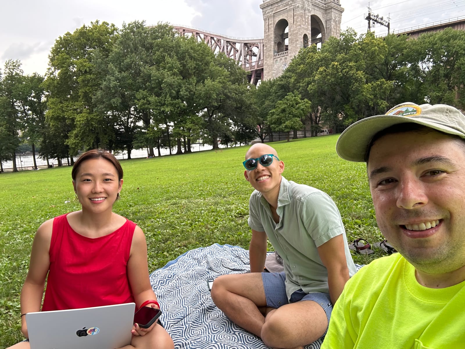 Three of us sitting on a blanket on the grass of Astoria Park's South Hill, laptop open, smiling at the camera, with the Hell Gate Bridge rising over the trees behind us.